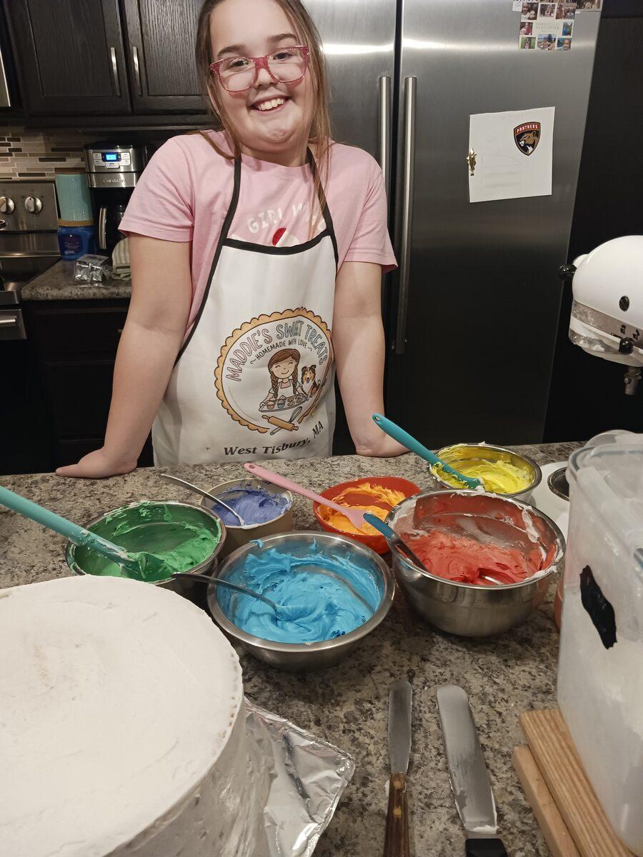Maddie in her Maddie's Sweet Treats apron, mixing bowls of rainbow frosting